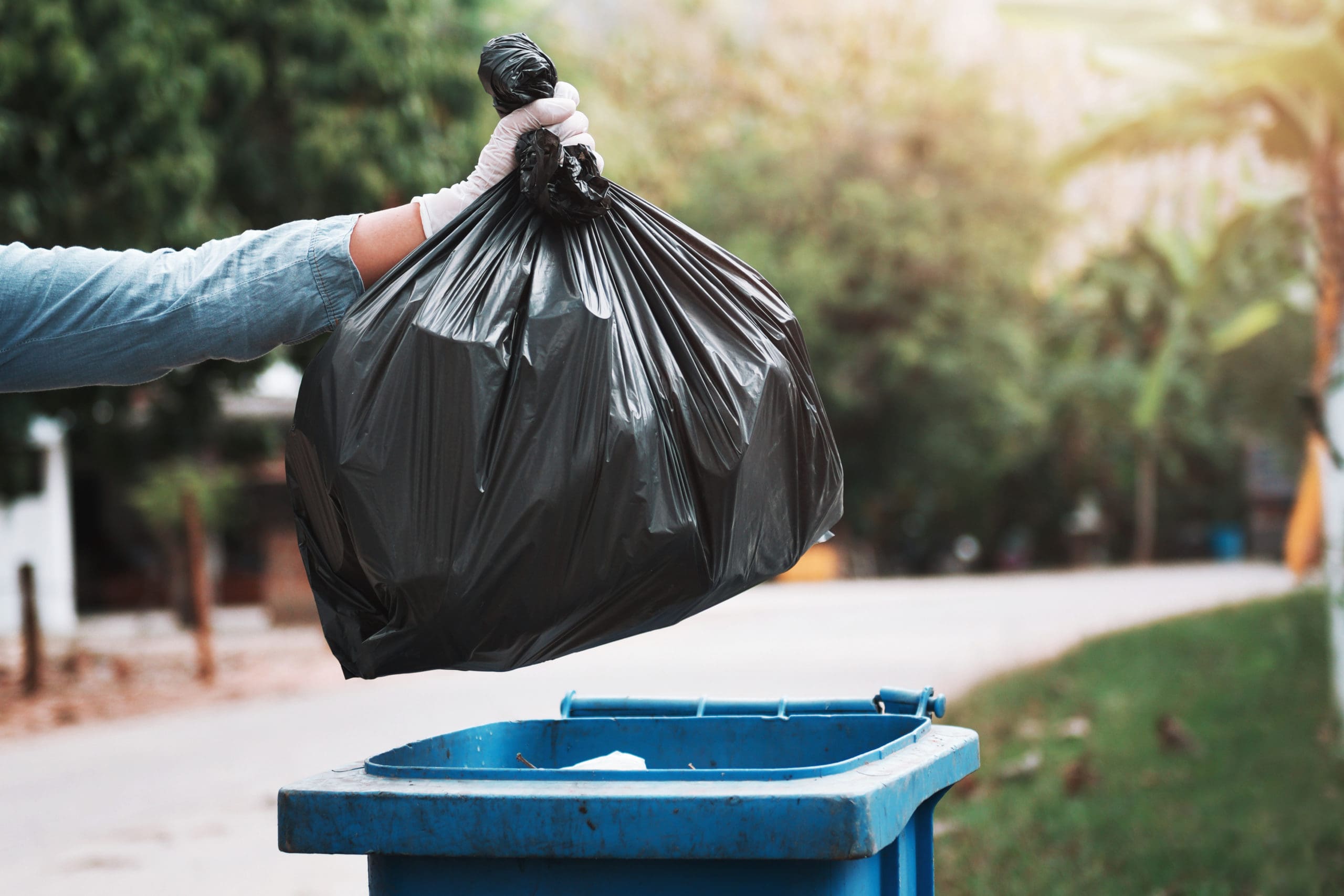 gloved hand holding black trash bag over a garbage bin for waste management collection