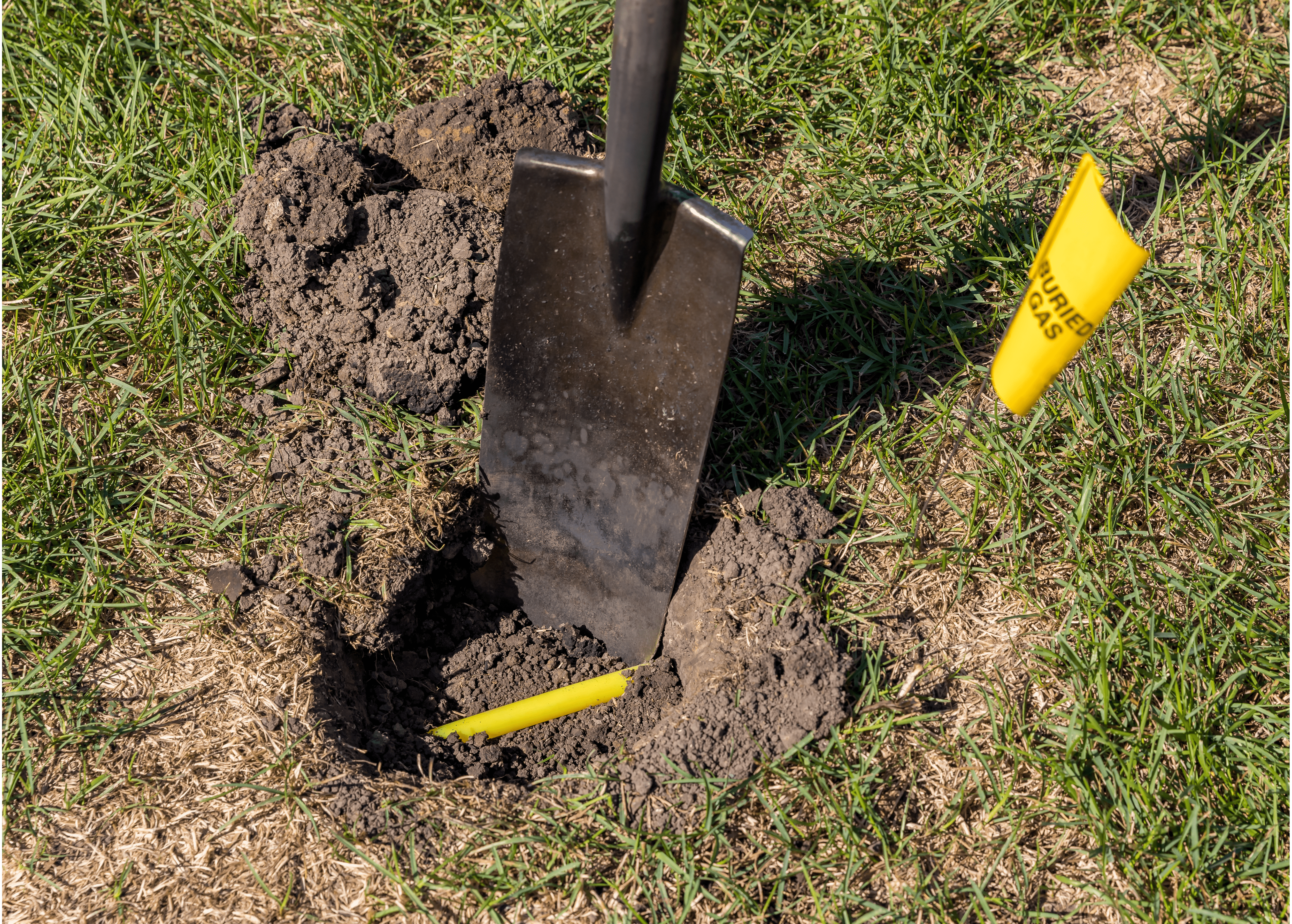 Hole in yard with yellow plastic natural gas utility lines buried in ground, shovel and with natural gas warning flag