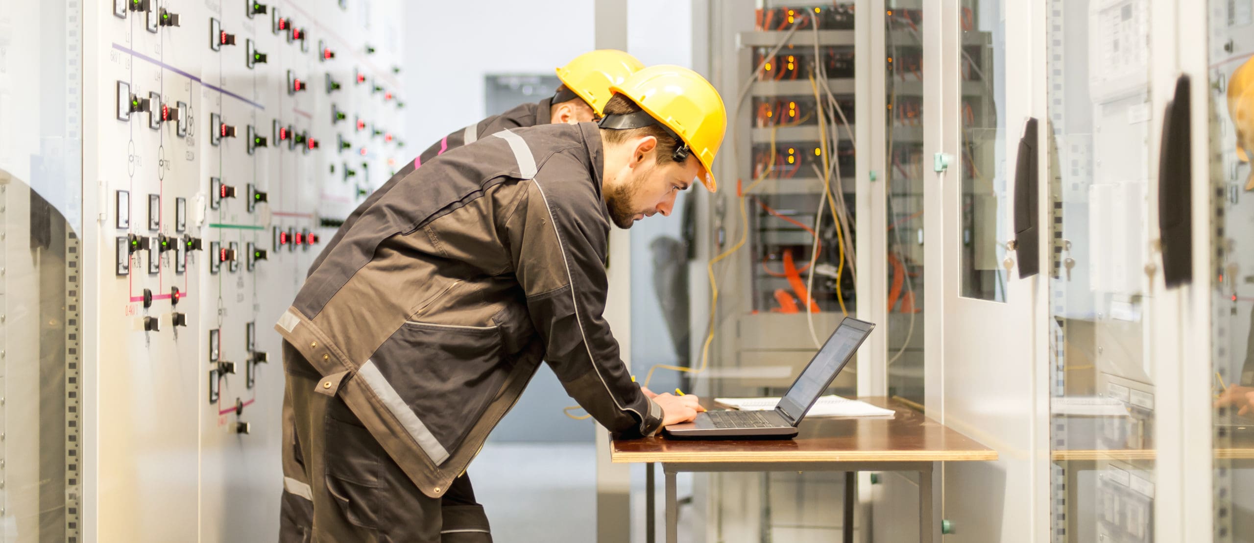 Two maintenance engineers inspect relay protection system with laptop computer. Bay control unit. Medium voltage switchgear