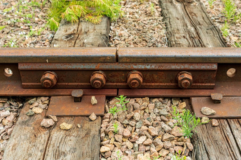 Detailed close up of a joint of an old railway track, with large rusty bolts and a large nail securing it to the wooden brace