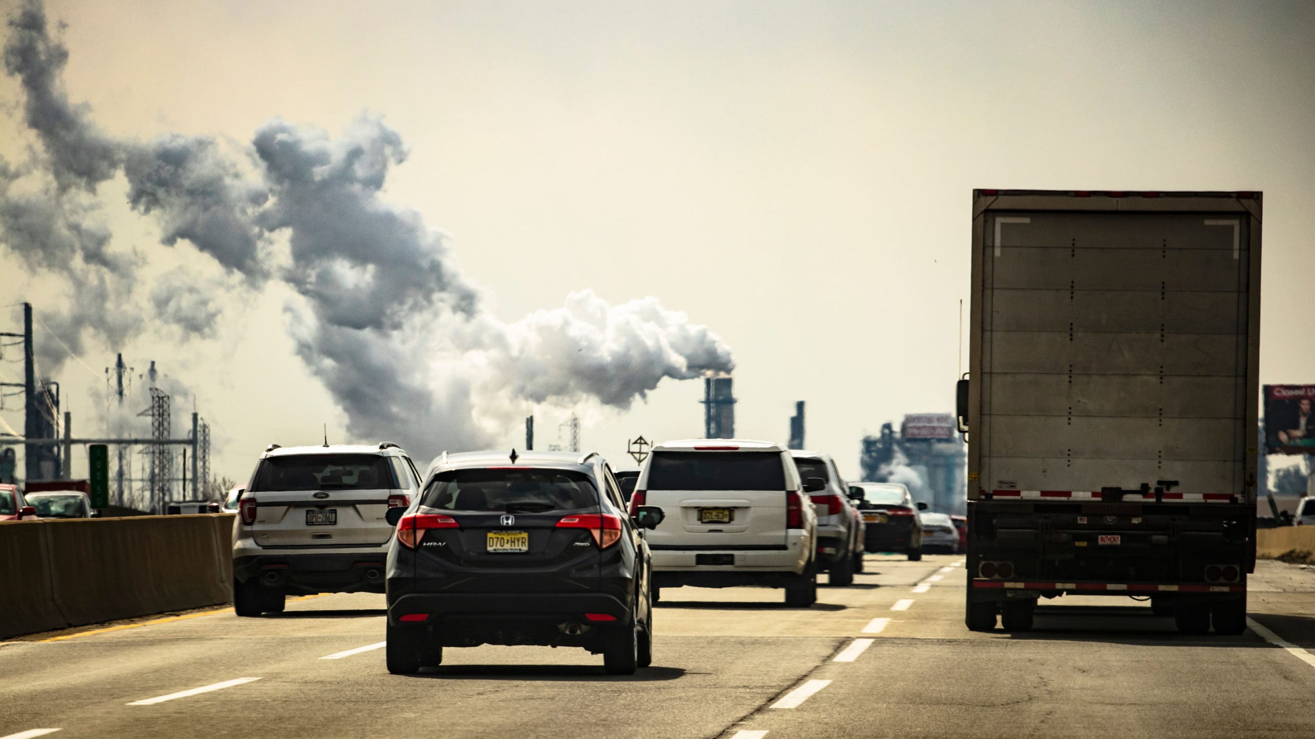 Smoggy view of cars and trucks on highway, with smoke billowing from factory in the background.